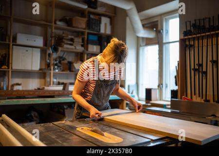 Fokussierte Zimmermannin bereitet Holzplatten für die Arbeitssäge vor, während Parameter in der Werkstatt gemessen werden. Stockfoto