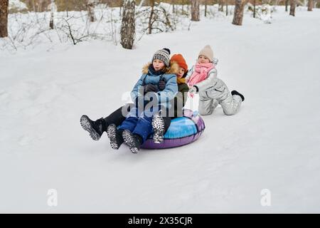 Zwei fröhliche, liebevolle Kinder rutschen auf Schneeröhren bergab, während das süße Mädchen in Winterkleidung sie beim Spielen im Winterwald drückt Stockfoto