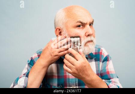 Alter grauer Mann Schnitt Bär mit Haarschneider. Porträt des Barbiers halten alte Rasiermesser Haarschneider, vintage Friseur, Rasieren. Alter bärtiger Mann. Vintage Stockfoto