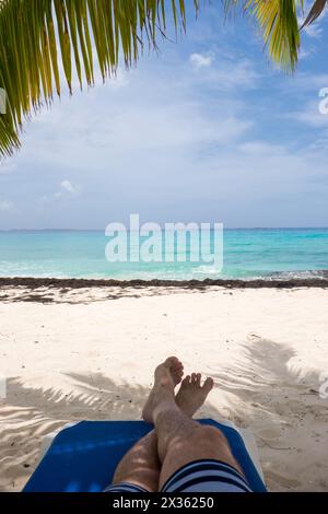 Ein Mann liegt auf einem blauen Liegestuhl mit den Füßen auf dem Sand. Der Strand ist ruhig und friedlich, mit dem Meer im Hintergrund. Der Mann hat Spaß Stockfoto