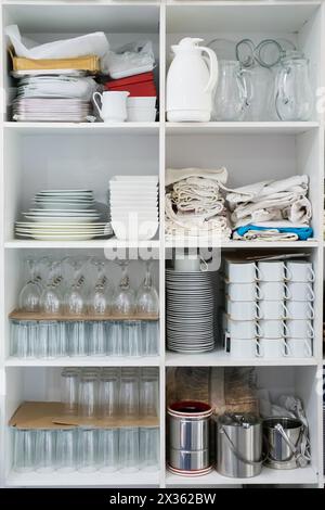 Ein weißer Schrank mit vielen Geschirr und Gläsern in den Regalen. Der Schrank ist voller verschiedener Gegenstände, darunter Tassen, Teller und Schüsseln. Szene ist unordentlich Stockfoto