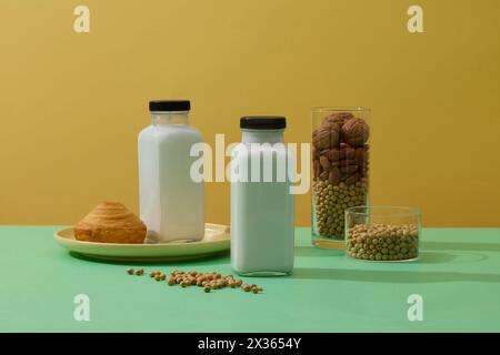 Zwei leere Flaschen mit frischer Milch, dekoriert mit einem Kuchen und Gläsern Sojabohnen, Mandel und Walnüssen. Gelber Hintergrund. Gesunde Bio-Lebensmittel Conce Stockfoto