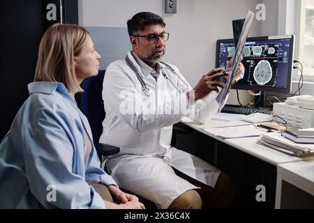 Birazialer Röntgenassistent, der eine Röntgenaufnahme des Gehirns bei reifer kaukasischer Patientin nach CT-Verfahren zeigt, Kopierraum Stockfoto