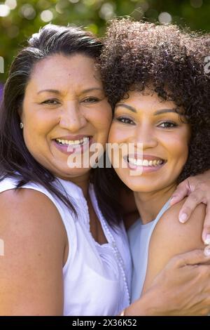 Reife, birassische Mutter umarmt ihre kleine Tochter, beide lächeln im Garten Stockfoto