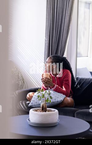 Eine junge afroamerikanische Frau sitzt auf der Couch, hält einen Becher und einen Kopierraum Stockfoto