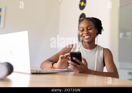Afroamerikanische junge Frau, die Ohrhörer trägt, zu Hause ein Smartphone und einen Laptop hält Stockfoto
