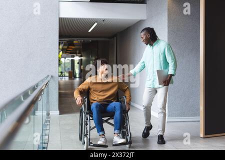 Afroamerikaner läuft neben lächelndem birassischen Mann im Rollstuhl in einem Geschäftsbüro Stockfoto