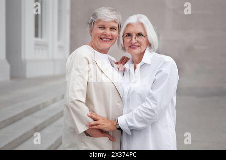 Porträt schöner lächelnder älterer Frauen mit grauen Haaren, die in stilvoller weißer Kleidung gekleidet sind, reife weibliche Models zusammen auf der Stadt Stockfoto