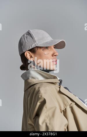 Ein junger, tätowierter Mann posiert selbstbewusst in Trenchcoat, Hut und Jacke vor grauem Studio-Hintergrund. Stockfoto