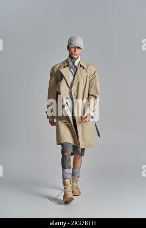 Ein stilvoller, tätowierter junger Mann im Trenchcoat posiert selbstbewusst in einem Studio vor grauem Hintergrund. Stockfoto