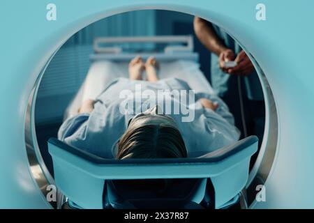 Reife Kaukasierin im blauen Kleid auf CT-Scannerbett in moderner Klinik liegend, unerkennbarer Arzt, der Einstellungen anpasst Stockfoto