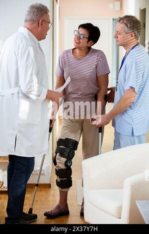 Eine ältere Patientin im Gespräch mit Ärzten im Kliniksaal. Frau mit Bondage, medizinischer Schiene, Kniestütze am Bein, Kommunikation mit dem behandelnden Arzt in einem Krankenhaus. Unterzeichnen von Versicherungspapieren Stockfoto