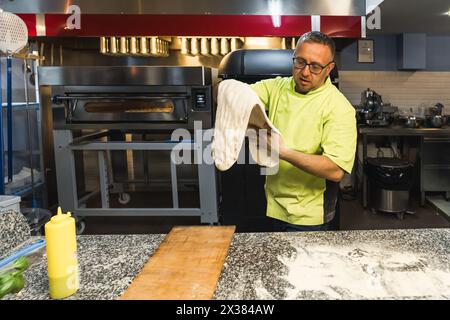 Professioneller Koch in Uniform hält einen riesigen flachen Pizzateig in der Restaurantküche. Hochwertige Fotos Stockfoto