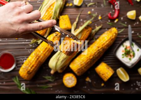 Küchenzange mit gegrilltem Mais auf den Kolbenstücken in kaukasischen Händen auf Küche Holztisch Hintergrund Stockfoto