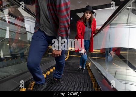 Junge lateinamerikanische Frau (23) geht die Rolltreppe in einem Einkaufszentrum hinauf. Sie trägt einen schwarzen Hut und einen roten Pullover. Wander- und Freizeitkonzept. Stockfoto