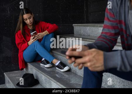 Junge Frau (23) Lateinamerikanerin, die auf der Treppe sitzt und ihr Handy überprüft. Selektiver Fokus auf sie. Technologie- und Bruderschaftskonzept Stockfoto