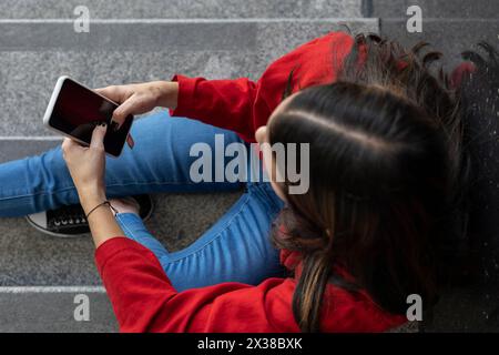 Junge (23) nicht erkennbare lateinamerikanische Frau (23), die auf der Treppe sitzt und ihr Handy überprüft. Blick von oben. Technologiekonzept. Stockfoto
