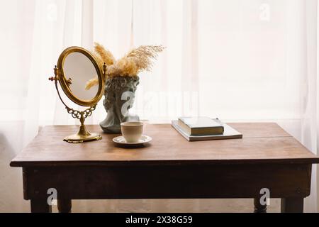 Eleganter Vintage-Spiegel und Büstenvase mit Pampas-Gras neben einer Kaffeetasse auf einem Holztisch Stockfoto