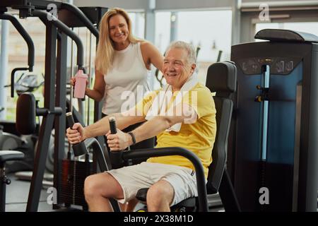 Aktive Seniorin, mit Wasserschlacht, spricht mit Mann, macht Handübungen, hebt Gewichte in einem modernen Fitnessstudio. Stockfoto