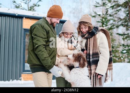 Mittelgroße Aufnahme eines kaukasischen Mannes, einer Frau und ihrer Tochter, die stilvolle Winterkleidung tragen und mit ihrem Hund im Freien spielen Stockfoto