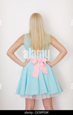 Junge Blondine im Kleid mit Schleife posiert in der Nähe der weißen Wand im Studio, Rückansicht Stockfoto