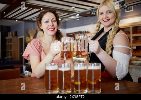 Zwei lächelnde Frauen klingen Glasbecher mit Bier über dem Tisch. Stockfoto