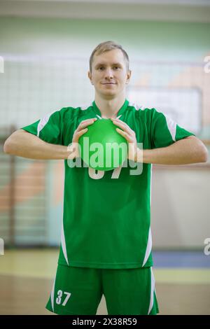 Mann in grüner Sportuniform hält Volleyballball und lächelt im Fitnessstudio Stockfoto