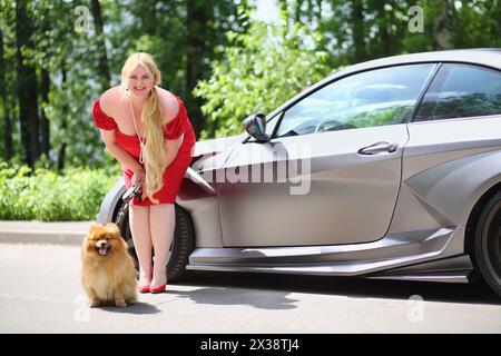 MOSKAU – 19. Juni 2016: Lächelnde Blondine (mit Modellfreigabe) mit langen Haaren in rotem Kleid steht mit einem kleinen Hund in der Nähe des Sportwagens nach vorne gelehnt Stockfoto