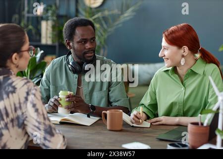 Porträt eines erwachsenen afroamerikanischen Mannes, der mit Kollegen spricht, die an einem Meeting-Tisch in einem modernen Büro mit grünen Pflanzen sitzen Stockfoto