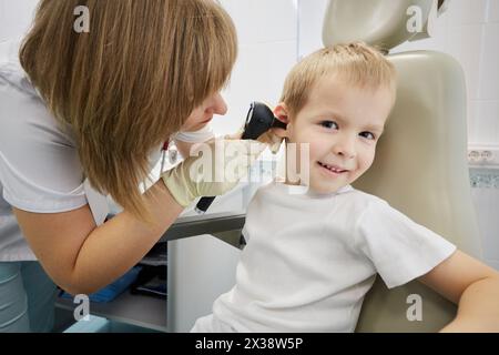 Weibliche Ärztin untersucht das Ohr des lächelnden Jungen, der im medizinischen Sessel sitzt. Stockfoto