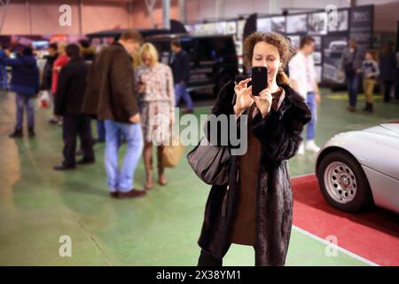 Glückliche hübsche Frau im Pelzmantel schießt telefonisch bei der Ausstellung Stockfoto