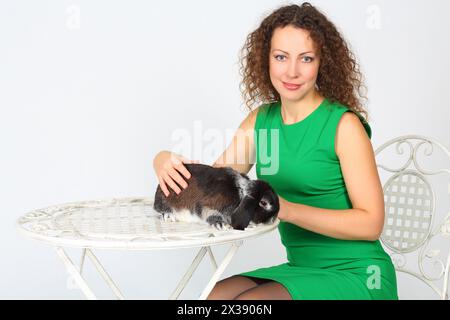 Hübsche Frau in Grün hält lustiges Kaninchen am Tisch im weißen Studio Stockfoto