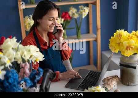 Blumengeschäft, Inhaber eines Blumengeschäfts. Frau, die Handy und Laptop benutzt, um Bestellungen für ihren Blumenladen anzunehmen. Hispanischer Gärtner nimmt Kunden Stockfoto