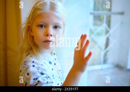 Ein hübsches blondes Mädchen steht neben dem Fenster im Zimmer und schaut in die Kamera Stockfoto