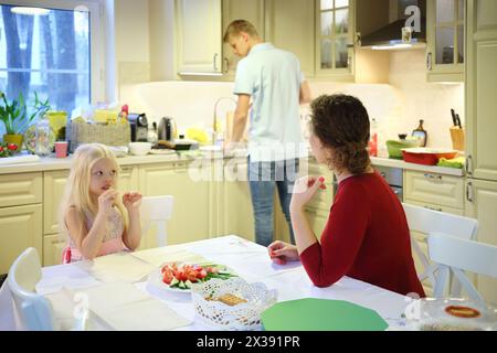 Hübsche kleine Mädchen und Frauen sitzen am Tisch und der Mann steht in der Küche weg, konzentrieren sich auf die Frau Stockfoto