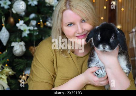 Die blonde Frau sitzt und hält den Hasen neben dem weihnachtsbaum in einem gemütlichen Zimmer Stockfoto