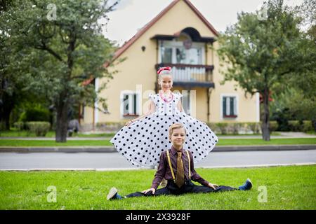 Der lächelnde Junge sitzt auf der Seite, und das Mädchen im gepunkteten Kleid steht hinter ihm auf dem Rasen vor dem zweistöckigen Haus. Stockfoto