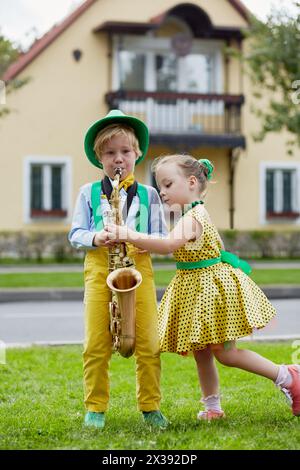 Der kleine Junge im Tanzanzug spielt Saxophon und das kleine Mädchen tanzt auf dem Rasen vor einem zweistöckigen Haus. Stockfoto