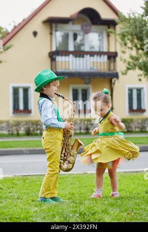 Der kleine Junge im Tanzanzug spielt Saxophon und das kleine Mädchen tanzt auf dem Rasen vor einem zweistöckigen Haus. Stockfoto