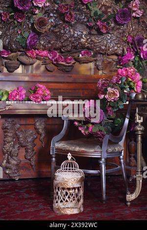 Altes Holz-Klavier, Stuhl und Vogelkäfig zwischen Rosen in stilvollem Studio Stockfoto