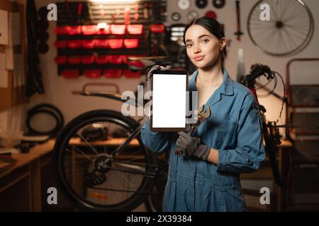 Junge Frau repariert Fahrrad in der Werkstatt, hält ein digitales Tablet mit Mockup-Bild Stockfoto