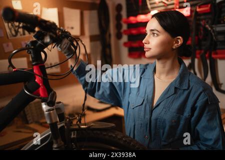 Fahrradmechaniker, der das Fahrrad in der Werkstatt oder in der Garage überprüft. Junge fokussierte kaukasische Frau. Fahrradservice, Reparatur und Upgrade-Konzept. Garageninnenraum Stockfoto