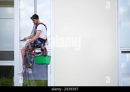 Der junge Mann hängt an einem Sicherheitsseil und reinigt das Fenster eines hohen Gebäudes Stockfoto