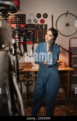 Frau Mechanikerin, die telefoniert, um Hilfe bei der Reparatur eines beschädigten Motorrads bittet, in Garage oder Werkstatt steht. Telefonanruf mit Fahrradingenieur Stockfoto