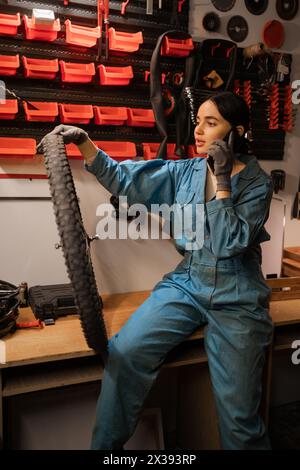Schöne brünette Frau in lässiger Kleidung, die auf dem Handy spricht, in der Fahrradwerkstatt oder in der Garage sitzt Stockfoto