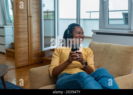 Ruhe- Und Entspannungskonzept. Porträt einer lächelnden Afroamerikanerin, die ein Mobiltelefon mit schnurlosem Headset benutzt und Musik online hört, fröhlich Casua Stockfoto