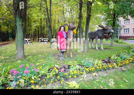 Glückliche Frau im Mantel posiert mit Holzhirschen in der Nähe von Elchen im Herbstpark Stockfoto