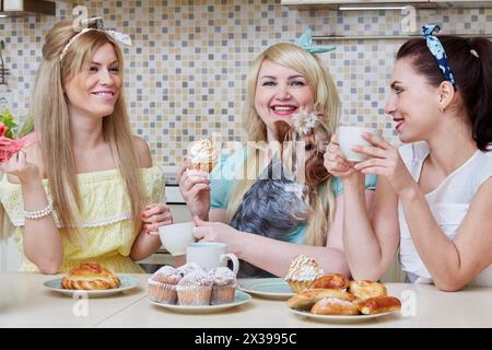 Drei junge lächelnde Frauen und ein kleiner Hund sitzen am Tisch mit gebackenen Süßigkeiten in der Küche. Stockfoto