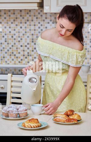 Eine junge Frau in gelbem Kleid gießt kochendes Wasser aus dem Wasserkocher in eine Tasse auf dem Tisch in der Küche. Stockfoto