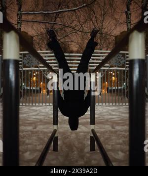 Mann kopfüber auf parallelen Bars auf dem Sportspielplatz im Hof in der Winternacht, Blick nach hinten Stockfoto
